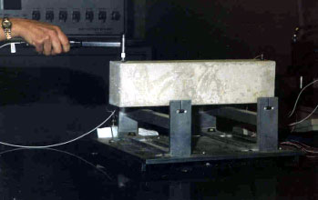 The picture shows an operator testing a concrete prism using the impact resonance frequency apparatus, which consists of: the support stand for specimen; the impact hammer, which is made of metal and has a spherical shape; and an accelerometer, which is attached to the concrete beam and is connected to the six-channel connector box. The apparatus is used in the impact resonance method (A S T M C 215).