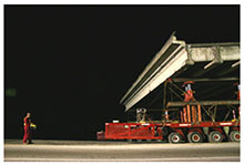 Self-propelled modular transporters (SPMTs) carry a bridge span down a road. The operator of the computer that controls the SPMT movement is walking behind the SPMTs.