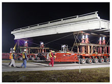 Self-propelled modular transporters (SPMTs) carry a bridge span down a road. The operator of the computer that controls the SPMT movement is walking behind the SPMTs, along with three other construction workers monitoring progress.
