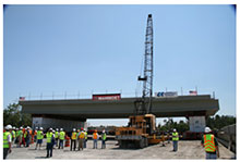 Self-propelled modular transporters carry a bridge span up a road to the bridge site. A couple dozen people with hard hats and safety vests are shown in the foreground as they observe the demonstration.