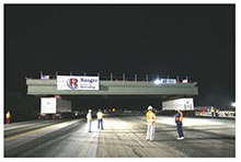 Self-propelled modular transporters carry a bridge span up a road to the bridge site during nighttime construction. Four construction workers with safety vests monitor progress.