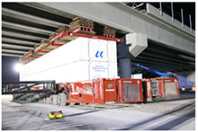 A close-up of a self-propelled modular transporter is shown underneath one end of a bridge span just after the SPMT lowered the span into position on the interior bridge support.