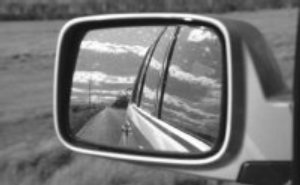Cameo: Reflection of passenger vehicle, highway, surrounding terrain, and sky as seen through a driver's side mirror.