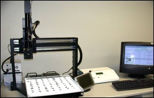 Figure 1. Photo. Original research Aggregate Imaging System equipment (AIMS1) (photo courtesy of Pine Instrument). On a table, at the right are a computer screen and keyboard; in the center are two pieces of equipment, a white, low device with a small display window and a small device similar to a small computer display panel. To the left is a white square tray containing pieces of aggregate in seven even rows. The tray is positioned on a platform at the base of and within a large open upright framework—one upright member at either side and a larger, flat bar across the top on which a tall black metal device is mounted that appears able to move back and forth over the tray. This device is plugged into a small box behind the apparatus.