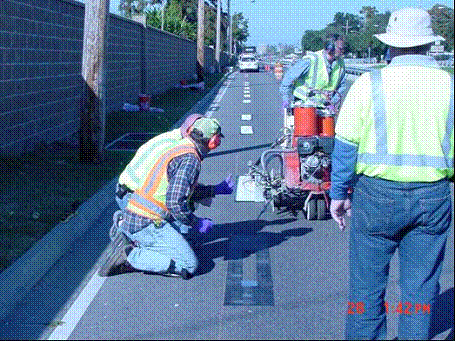 This image shows the installation crew during the test marking installation. One of the crew members install the markings with a self-propelled cart with a double drop system. Two of the crew members view the bead element drop from respective nozzles as the cart passes by, spraying the paint and dropping the beads and elements.