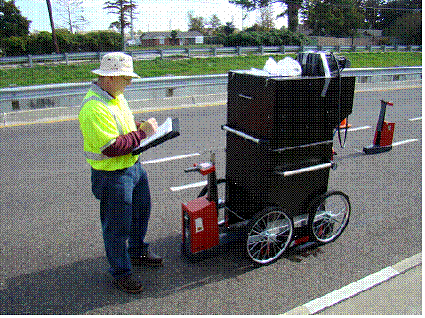 This picture shows a view of the rain simulator and a crew member noting the measured retroreflectivity from an LTL-X type handheld retroreflectometer placed directly underneath the portable rain simulator. The rain simulator is a stand-alone unit, which has four wheels that can be pushed by hand. It is approximately 5-feet high, 2-feet wide, and 2.5-feet long, and has a hollow section toward the bottom, where the portable retroreflectometer is placed.