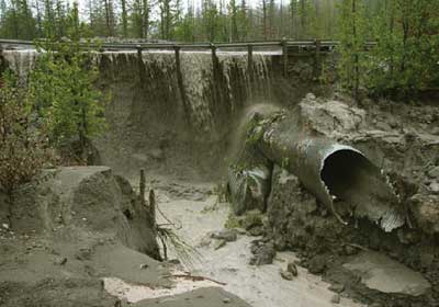 Photograph. A failed metal culvert is shown, with water cascading over the roadway above.