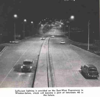 Photo of a highway.  Caption says 'Sufficient lighting is provided on the East-West Expressway in Winston-Salem, which will become a part of Interstate 40 in the future'.