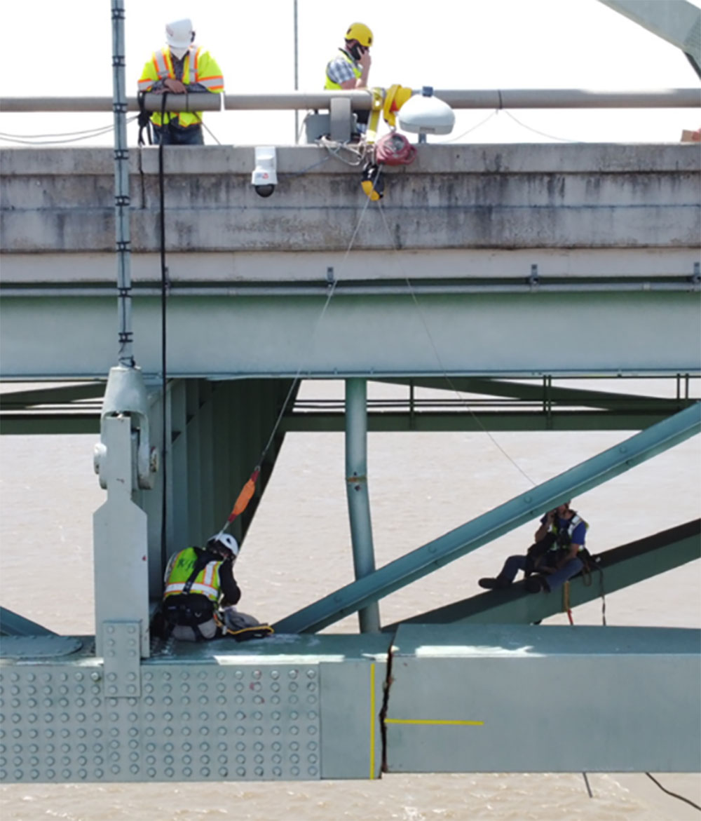 View of Hernando DeSoto bridge from side. A visible fracture going all the way through the beam is present on the lower support of the bridge. Four workers are present, two at road-level and two on beams underneath the bridge.