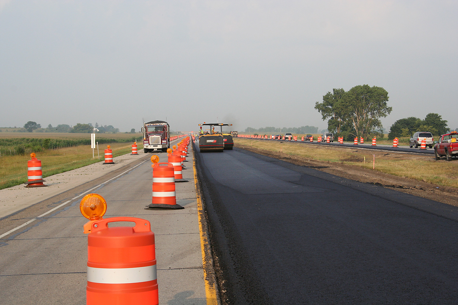 A divided four-lane highway where the two inside lanes are closed to traffic, separated by orange traffic drums. Traffic flows on the open lanes while construction equipment is present on one lane that has been overlayed with new asphalt.