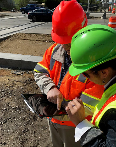 Man and woman wearing orange and yellow safety vests and hardhats standing at a construction site referring to information on a mobile tablet.