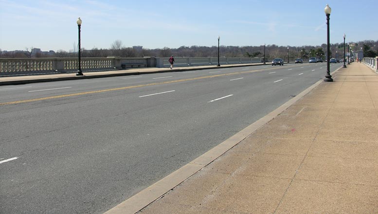 This slide shows an image of the six-lane bridge deck of the Arlington Memorial Bridge. There are three lanes in each direction, and there are sidewalks and concrete railings visible on each side of the bridge deck. There is an asphalt overlay on top of the concrete bridge deck. There are light posts on the sidewalk; vehicles on the road; and pedestrians on the sidewalks. There is a heading of “Description of Existing Bridge” and bulleted text above the image of the bridge deck. Trees and buildings can be seen in the distance beyond the bridge deck. The slide number is visible in the lower right corner.