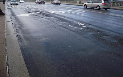 This slide shows four images identifying deterioration of the roadway surface (bridge deck) of the Arlington Memorial Bridge. The slide in the upper left shows the bumpy surface of the roadway (bridge deck), including ruts, spalls and asphalt patches. Bulges in the pavement are also visible from repeated repair/patching with asphalt. The image in the upper right shows a hole in the road way surface (bridge deck) where the entire asphalt overlay is missing and the concrete bridge deck below it is showing. Also visible is a dark area of asphalt where the roadway had been previously repaired/patched. The image in the lower left shows the sidewalk of the bridge; the sidewalk has large areas of deterioration and cracking It also shows where the deterioration in the sidewalk had been patched with asphalt but the patch also has deteriorated. The image in the lower right shows a pothole in the roadway surface (bridge deck). The asphalt roadway surface also has ruts and there are areas on the road surface that have been previously repaired/patched. The slide number is visible in the lower right corner.