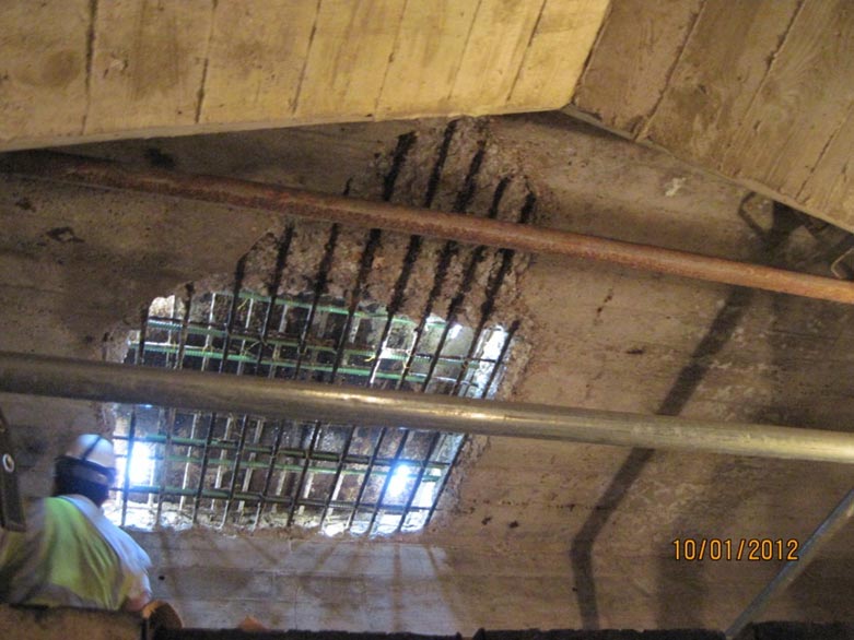 This slide shows an image of the underside of the deck of the Arlington Memorial Bridge taken on October 1, 2012. The photograph is looking up at the bridge deck from below, and shows the rectangular hole in the deck and reinforced bars. Additional corroded reinforcing bars are visible adjacent to the hole in the bridge deck, and the concrete cover has been lost in the area of the corroded reinforcing bars. A worker is also in the photograph, wearing a bright yellow and white safety shirt and a hardhat. The image also shows pipes/conduits carrying utilities under the bridge deck. The slide number is visible in the lower right corner.