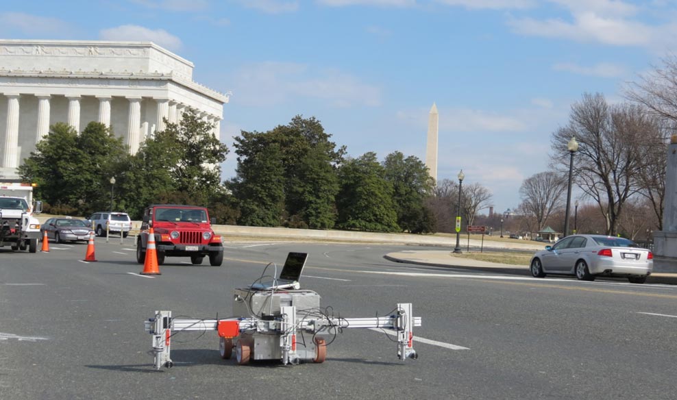 The tool is comprised of a silver box on four small wheels. It has a horizontal supporting bar on the front of it and that horizontal bar supports one vertical arm on each side. The vertical arm on each side contains the impact echo impulse and measuring devices. The tool is located on the bridge deck of the Arlington Memorial Bridge at the end of the bridge near the Lincoln Memorial. The bridge deck contains an asphalt overlay (wearing surface). Behind the bridge deck and to the left of the IE tool are vehicles, traffic cones, and the Lincoln Memorial. Behind the bridge deck and to the right of the IE tool is a parked vehicle with the Washington Monument in the distance behind the some trees.