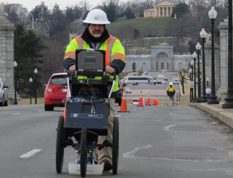 An image of a worker using ground penetrating radar (GPR) on the Arlington Memorial Bridge. Vehicles are seen on the bridge but the lane in which the worker is using the GPR equipment is closed and orange traffic cones separate the open lanes from the closed lane. In the background, portions of Arlington National Cemetery are seen.