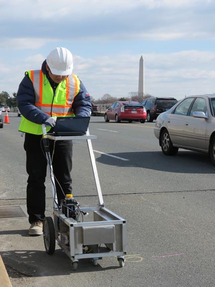The first photograph shows a worker wearing a safety vest and hard hat and using a tool to measure concrete quality using ultrasonic surface waves. The tool is contained in a push cart with wheels. The ultrasonic surface wave equipment is contained in a box at the bottom of the cart, while a computer is recording the data. The image includes a worker using the tool and the computer on a closed lane of the Arlington Memorial Bridge. Vehicles are seen on the roadway and the Washington Monument can be seen in the background.