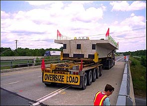 The photograph on the left features a tractor trailer on top of a bridge with a large concrete structure on the bed of the truck. The words “Oversize Load” appear on a sign on the back of the trailer, and orange flags appear on the concrete structure and on the truck. A worker wearing an orange and yellow safety vest appears in the lower right portion of the photograph.
