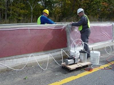 Figure A4b.F3. Photo. Typical vacuum impregnation setup. Photo shows the side of a concrete barrier with extensive cracking covered by a red plastic mesh taped to it. A spiral plastic tube is adhered to the top of the mesh, near the top of the barrier by the contractor. The vacuum has been turned on, and the lithium is being vacuumed across the barrier, first starting in the lower right corner and radiating out towards the center of the barrier.
