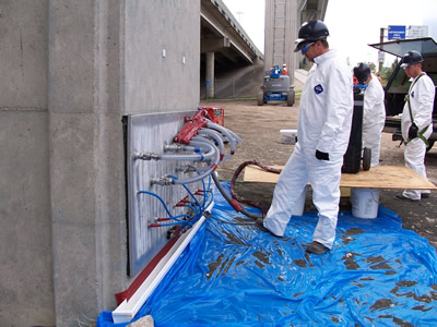 Figure A4b.F4. Photo. Vacuum impregnation treatment in Houston on a concrete column. A metal slab is being applied to the side of a concrete column, and manages to stay on through a vacuum process. Several plastic clear tubes, taped together, are adhered to the metal slab, with one main grey tube attached to a compressor that let the solution penetrate the structure.