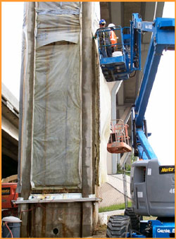 This image shows a concrete bridge column covered with plastic sheeting on all four sides. Two men are being lifted by a mechanical crane about 20-25 feet above ground. 