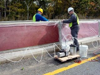 This image shows a highway concrete barrier being treated using a vacuum impregnation method. red plastic mesh covers the sides of the barriers, and a clear plastic cover is being applied by two men.