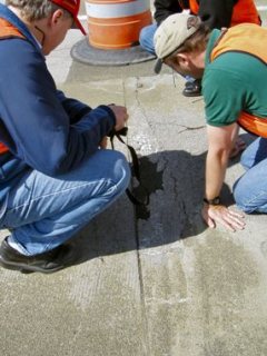 This image shows two men looking over a concrete pavement near a transverse joint. There is an area near the jont exhibiting severe damage, and has been surfaced with black material.