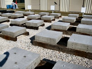 This image shows a close up view of the bridge deck concrete samples. Several large square shaped concrete samples are being supported by wooden beams on top of a gravel area.