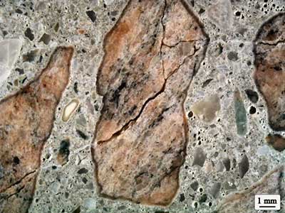 This image shows a close-up of a concrete sample. In the middle of the sample is a granitic gneiss aggregate, salmon in color, with several cracks running through it. One large crack extends to the surrounding aggregates, running through them as well.