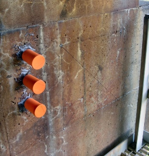 This image shows a monitoring section on the surface of a cracked concrete structure. Three orange plastic cylindrical cups are attached to the wall vertically, one under the other. To the right of the cups, a grid is drawn out with three steel pins at the top left, top right, and bottom left corners of the grid.