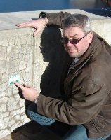 Photograph of editor, Michael Thomas, kneeling next to a concrete barrier exhibiting dark cracks and holding a crack comparator card.