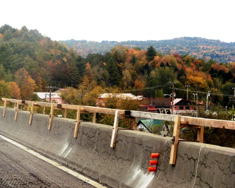 This image shows a concrete bridge barrier wall with extensive cracking running parallel to the bridge deck. Three small orange plastic containers are seen attached to the side of the barrier wall, and a constructed wooden guardrail is located on top of the wall, running the span of the wall.