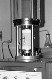 Close-up view of Shedworks Simple Performance Test device. The glass chamber hood is lifted, exhibiting the asphalt specimen loaded and ready for testing inside. The asphalt core specimen is enclosed in a rubber-plastic sheath. A metal transducer is attached, snapped into two smaller metal brackets, on the side of the asphalt specimen.