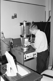 View of man placing asphalt core specimen (wrapped in rubber-plastic sheath) into glass chamber and onto the loading platen inside simple performance test (Shedworks). In the foreground, a view of the accompanying computer is shown.