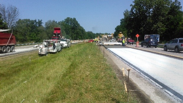 Concrete is being paved on a roadway. The concrete paving equipment is shown along with traffic control for the bypassing vehicles.