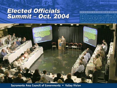 Elected Officials Summit - Oct. 2004. Large photo shows a meeting of people in a room with tiered seating in a u-configuration, and two projection screens and a dais in the open end.