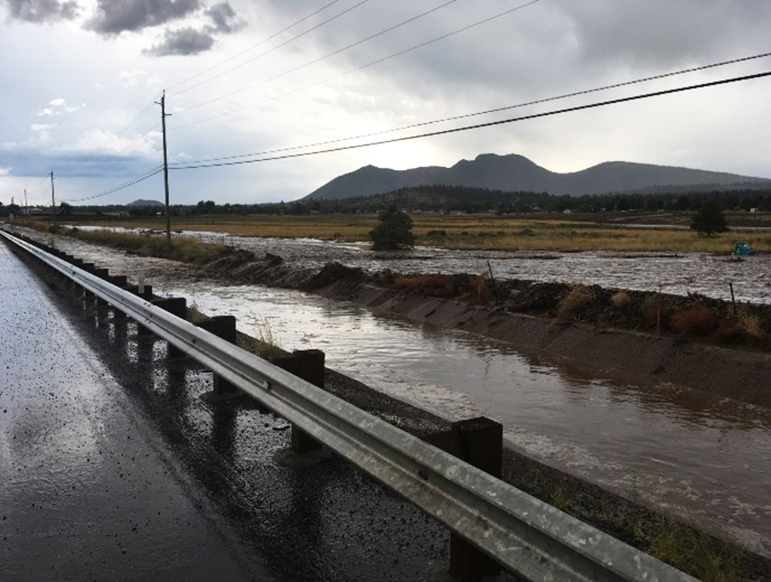 In 2018, the new concrete lined channels along US 89 directed floodwaters to the detention basin and protected the roadway and surrounding homes from major damage. A drainage ditch located next to a wet road and guardrail is filled with water.