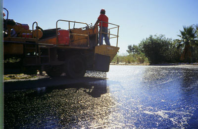 Figure 8. Photo. A chip seal is applied to an asphalt pavement. A worker can be seen standing on top of a spreader.