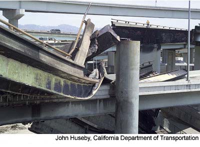 Figure 2. Photo. A close-up view of the damaged I-880 and I-580 connector roads. The concrete deck and steel girders are mangled and jutting out. A worker can be seen surveying the damage.