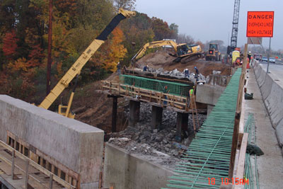 Figure 3. Photo. I-96 Superstructure Replacement. A new bridge is under construction on I-96 in Michigan. A sign reads "DANGER. Overhead Lines. High Voltage." A crane and other construction equipment are visible. Five workers are on scaffolding in a pit next to the roadway. Two cars and a truck are on the roadway.