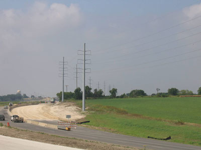 Figure 5. Photo. State Highway 130 Toll Road Project, Texas. Cars and trucks travel on the two left lanes of the roadway, while the right lanes are closed off as a work zone. A row of seven power lines are to the right of the work zone.
