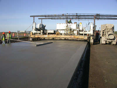 Photo. A shot of concrete paving. Paving equipment is in the background. Three workers with hard hats are visible.