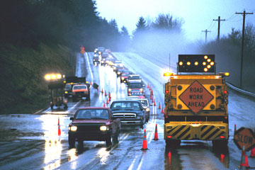 Figure 6. Photo. Cars travel through a work zone in the rain. A lane is blocked off, with a sign that reads "Road Work Ahead" and an arrow pointing right on top of the sign.