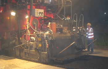 Figure 3. Photo. A worker stands next to a paver that is applying an ultra-thin hot-mix asphalt overlay as a preventive maintenance treatment on I-20 in Louisiana.
