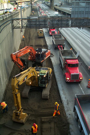 Figure 8. Photo. Concrete pavement construction on a highway. Dump trucks, bulldozers, and construction workers are visible.