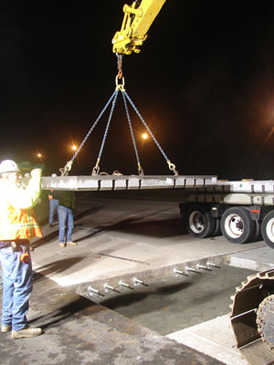 Figure 7. Photo. A precast concrete pavement slab is lifted into place by a crane. A worker is to the left of the slab.