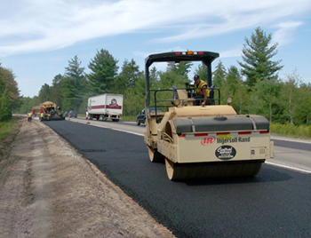 Figure 1. Photo. The Michigan Department of Transportation performs asphalt paving on a two-lane road on M-115 in Clare County, Michigan. A paver with an operator is visible in the foreground, with traffic in the background traveling in the other lane.