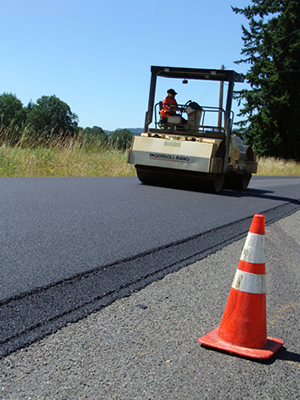Figure 7. Photo. A worker operates paving equipment on an asphalt roadway. A traffic cone is in the foreground.