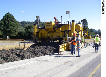 Concrete pavement under construction. Paving equipment and five workmen are visible.