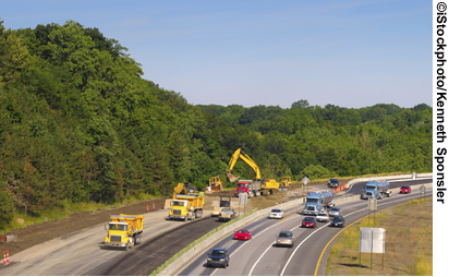 A view of a highway work zone. Traffic in the right hand lanes of the highway is moving, while the left hand side is closed off to traffic. Trucks and other construction equipment are visible.